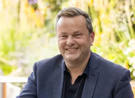 Simon Miles, wearing a dark suit and smiling at the camera whilst sitting at a table outside